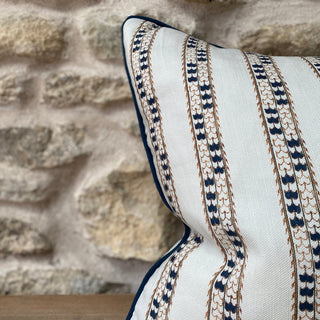 A close-up of the corner of a cushion, set against an exposed stone wall. The cushion is a natural colour with a detailed dark blue patterned stripe and dark blue piping.
