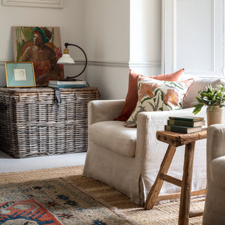 Pink square suzani flower cushion with green, yellow and grey leaves and details on a cream background. The pillow has pink piping around the edge and is sat on a natural linen chair in front of a pink velvet cushion. There is a patterned rug & art.