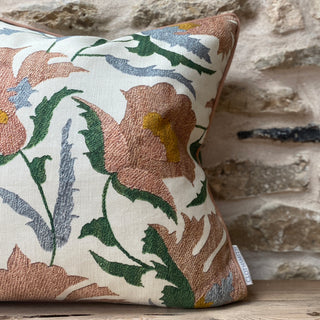 A pink square suzani flower cushion, with green, yellow and grey leaves and details on a cream background. The pillow has pink piping around the edge and is sat on wood against a stone wall.