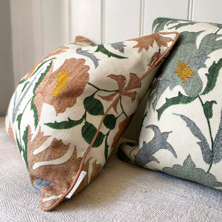 Two square suzani flower cushions, one in pink and the other in blue green. Both have green, yellow and grey leaves and details on a cream background. The pillows have piping around the edge in the main colour and are sat against panelling.