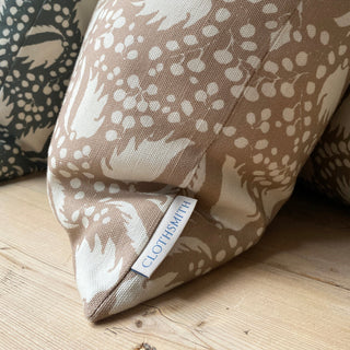 The picture shows the bottom corner of a square cushion placed on wooden table. The cushion features a blush pink background with an intricate cream pattern of leaf-like shapes and clusters of small dots. The pattern is dynamic and organic, giving it an elegant appearance.