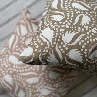 Close-up of a gold cushion with an off white floral and leaf design, with a blush pink cushion in the same pattern out of focus in the background. Both are placed on a rug with subtle grey and white stripes.
