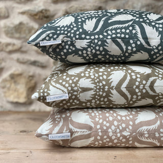 A pile of three cushions, all with an off white leaf pattern. The bottom one has a blush pink background, the middle one gold and the top one green grey. They are sitting on a light wooden surface with an exposed stone wall out of focus in the background.
