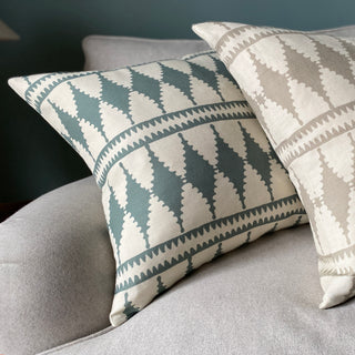 A close-up of two cushions are leaning against the arm of a grey wool sofa. Both cushions have a bold diamond pattern with cream background. The more prominent cushion is in green blue, and the other is in a natural colourway. The wall behind is a deep bluegreen.