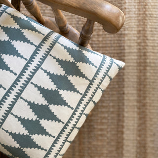 A close-up from above of the corner of a cushion with a bold diamond pattern in slate green and cream on a wooden armchair. Out of focus on the floor is a striped jute rug.