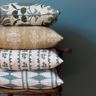 A pile of four cushions sit to the left of the image on a wooden armchair, with a dark blue green wall behind. The bottom cushion has a bold diamond pattern in cream and blue green, the next is cream with a subtle blue and beige stripe pattern, the one above is in a hay yellow with a delicate striped pattern in cream, and the top cushion is cream with a distinctive floral pattern in green blue.