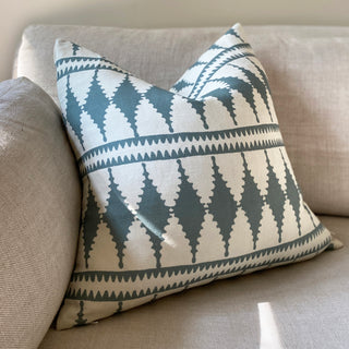 A picture of a cushion leant against the armrest of a natural linen sofa in dappled sunlight. The cushion has a bold diamond pattern in cream and slate green.
