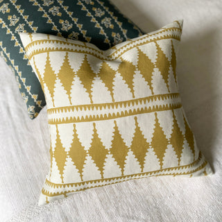 Taken from above, this is a picture of a square cushion with a bold diamond shaped pattern in ochre and cream. It is lying on a cream throw and leaning against another cushion in slate green with a delicate striped pattern.