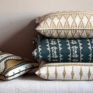 A pile of three cushions set to the right of the image on a cream bench, with a smaller cushion lying to the left side. All cushions are patterned in ochre, cream and slate green.
