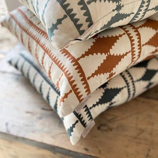 A close up of the corners of pile of three cushions, all in the same bold diamond pattern. All with cream backgrounds, the cushions are in slate green, terracotta and green blue. They are sitting on a wooden trunk.