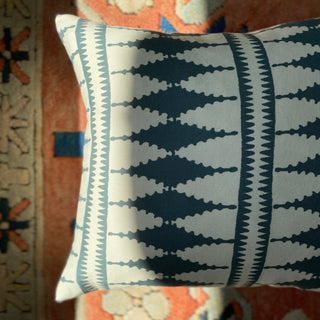 A close-up from above of a square cushion, with sunlight hitting the side of it. The cushion has a bold diamond pattern in slate green and cream, and is lying on a terracotta patterned rug.
