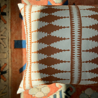 A close-up from above of a square cushion, with sunlight hitting the side of it. The cushion has a bold diamond pattern in terracotta and cream, and is lying on a terracotta patterned rug.