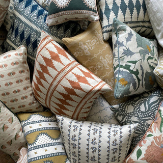 A close-up of heap of of patterned cushions, all in different colours and patterns. The most prominent cushion has a bold diamond pattern in terracotta and cream.
