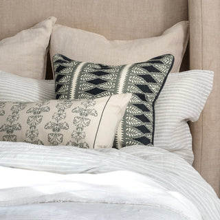 A close-up of a bed with a tall linen headboard. The image shows striped bed linen in soft grey and white stripes, with two feature cushions leant against the pillows. The cushions are both in cream and dark blue colour ways, with different detailed patterns. There is a white throw in the foreground.