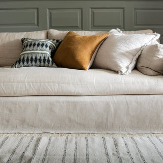 A cropped image of a natural linen sofa with a grey green panelled wall behind it and a cream striped rug on the floor infront. On the sofa there are three square cushions, on in plain natural linen, one in a  burnt orange velvet and the other in a dark blue and cream patterned print.