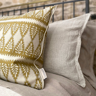 A close up of a bed with natural striped pillows and a black cast iron bed frame. A patterned square cushion sits in front of the pillow, with a detailed scallop pattern, in an ochre and cream colour way.