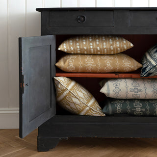 The picture shows a close up of half a black wooden cupboard with open doors. Inside the cupboard there are a piles of patterned cushions in ochre, hay yellow and slate green. There is a cream panelled wall behind and a light wooden floor.