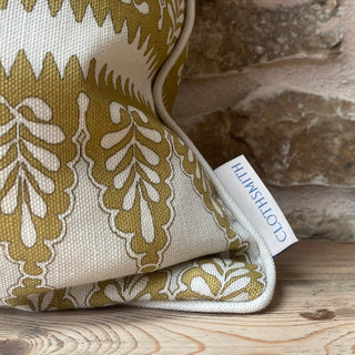 A detail shot of the corner of a patterned cushion in ochre and cream. There is cream piping on the cushion and a branded Clothsmith label. It sits in front of an exposed stone wall.