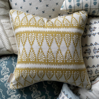 A square patterned cushion taken from above sits on top of a pile of other patterned cushions. The cushion is in ochre and cream colours, and has a detailed zig zag pattern.