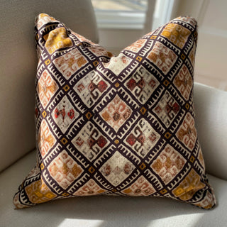 A cushion with a geometric pattern in black, pink and ochre sits on a cream textured armchair, with sunlight falling across it creating shadows.