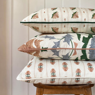A pile of three cushions sitting on a wooden stool. All of the cushions are patterned in varying designs, all in cream, pink and green colourways. There is a cream panelled wall behind.
