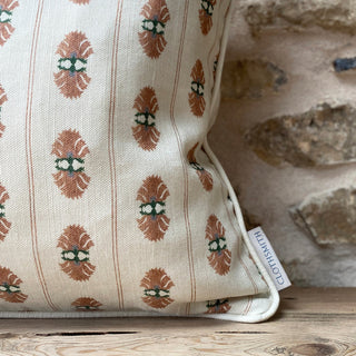 A close up of the corner of a square patterned cushion with a pink floral stripe on a cream background. The branded Clothsmith label can be seen in the bottom left corner. The cushion is leant against an exposed stone wall.