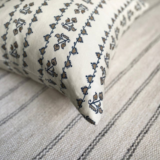 A close up of the corner of a cushion on a cream rug with grey stripes. The cushion is cream with a detailed dark blue patterned stripe.