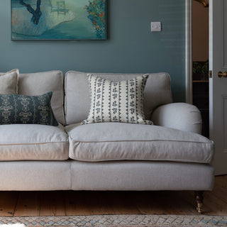 A light grey wool sofa sits next to a half open door, against a deep green blue wall. The sofa has cushions on it, the most prominent cushion in cream with a detailed dark blue patterned stripe.