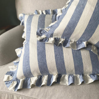 Two cushions with wide blue and white stripes are thrown onto a grey wool sofa. The cushions are frilled and only the corner of the cushion on top can be seen.