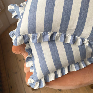 Two cushions with wide blue and white stripes are sat on a terracotta velvet armchair. The cushions are frilled and the chair sits on a natural wooden floor.