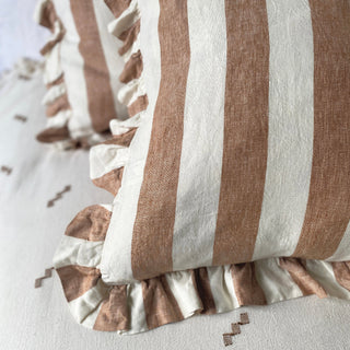 A close up of two linen cushions on a cream patterned bedspread. Both cushions have wide terracotta and white stripes with a frill. The cushion behind is out of focus.