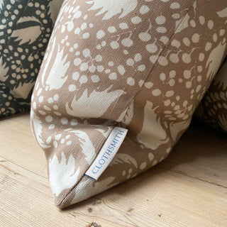 The picture shows the bottom corner of a square cushion placed on wooden table. The cushion features a blush pink background with an intricate cream pattern of leaf-like shapes and clusters of small dots. The pattern is dynamic and organic, giving it an elegant appearance. 