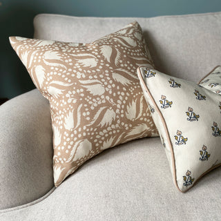  The picture shows two decorative cushions placed on light wool sofa. One of the cushions features a blush pink background with an intricate cream pattern of leaf-like shapes and clusters of small dots. The pattern is dynamic and organic, giving it an elegant appearance. The second cushion features small, delicate floral motifs in shades of blush pink and ochre on a natural background. This cushion also has blush pink piping along the edges.