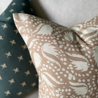 The picture shows the corner of a cushion placed on light wool sofa with another slate green patterned cushion behind it. The cushion features a blush pink background with an intricate cream pattern of leaf-like shapes and clusters of small dots. The pattern is dynamic and organic, giving it an elegant appearance. 