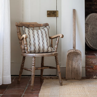 A natural striped square cushion sits on a wooden armchair in the corner of a dining room, with a tiled teracotta floor and jute rug underneath. An antique wooden shovel is leaning up against the wall next to the chair.