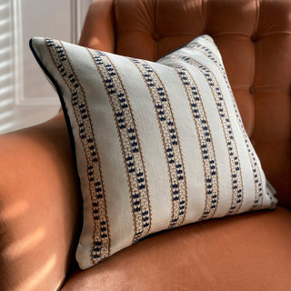 A close-up of a square cushion in a natural colour with detailed blue patterned stripes and blue piping sits on a terracotta velvet armchair with buttons. Light from the window shutters falls across the chair.