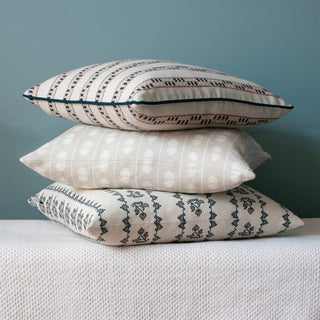 A pile of three cushions, sitting on a textured cream bench, set against a deep blue green wall. The cushions are all cream with slightly different patterned stripes in a colour palette of navy blue and natural.