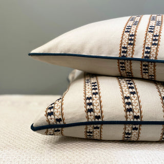 A close-up of the corner of two cushions, one on top of the other, sitting on a textured cream throw against a grey wall. The cushions are a natural colour, with a detailed pattern stripe in navy blue and navy blue piping.