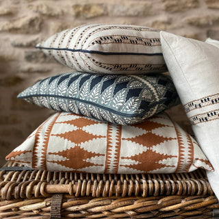 A pile of four cushions sitting on a wicker basket with an exposed stone wall behind. The bottom cushion has a bold diamond pattern in cream and terracotta, whilset the top cushions are a combination of patterns in natural and navy blue stripes. One of the cushions is leant up against the pile and is half out of the shot.