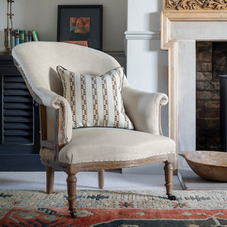 A natural linen armchair with wooden legs sits next to a fireplace with a black chest behind it and a patterned red rug beneath it. There is a large wooden bowl infront of the fireplace. There is a natural square cushion with detailed stripes in navy blue and matching piping sitting on the chair.