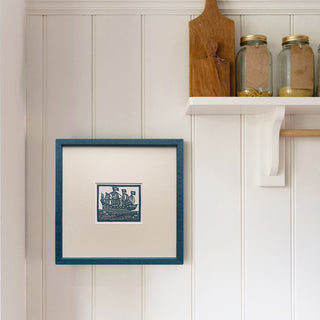 Framed teal blue artwork of a galleon ship on a wall next to a shelf with jars and a chopping board.