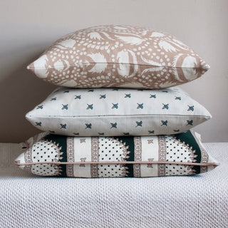 The picture shows three cushions piled on top of each other. The top cushion features a blush pink background with an intricate cream pattern of leaf-like shapes and clusters of small dots. The pattern is dynamic and organic. The second cushion is a natural colour and features small grey green ditsy flowers and is simpler in appearance. The bottom cushion features a botanical inspired design, covered in a rambling pattern in blush pink and slate green on a natural background.