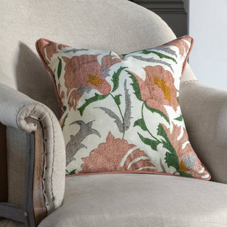 Pink square suzani flower cushion with green, yellow and grey leaves and details on a cream background. The pillow has pink piping around the edge and is sat on a natural linen chair with exposed wood back.