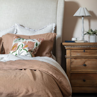 Pink square suzani flower cushion with green, yellow and grey leaves and details on a cream background. The pillow has pink piping around the edge and is laid on a natural linen bed with pink bedding. There is a side table to the right with a lamp.