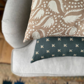 The picture shows the corner of a cushion from above placed on light wool sofa with another slate green patterned cushion below it. The cushion features a blush pink background with an intricate cream pattern of leaf-like shapes and clusters of small dots. The pattern is dynamic and organic, giving it an elegant appearance. 