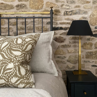 A square brown gold cushion with cream leaf pattern sits on cast iron black bed with natural striped bed linen. Behind it is an exposed stone wall and it has a gold lamp with black shade next to it.