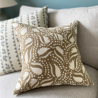 A sandy gold cushion with an off white floral and leaf pattern, placed on a light grey sofa, with another cushion featuring a subtle blue and white design in the background.