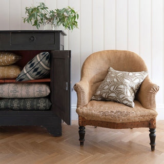 An antique French armchair with visible wear, featuring a cushion with an off white and gold leaf pattern. Next to it is a dark wooden cabinet with its door open, displaying patterned cushions. A vase with greenery sits atop the cabinet, and the setting features light wooden flooring and a white panelled wall.