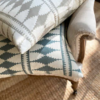 A close-up of two cushions lying on a natural linen armchair. Both have a bold diamond pattern, the bottom one in a blue green and cream colourway and the top in natural stone and cream. There is a jute rug out of focus on the floor beneath the chair.