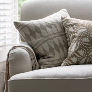 Two cushions on an oatmeal armchair with a throw hanging over the armrest. The more prominent  cushion is leant against the armrest, with a bold cream and natural diamond pattern. The other cushion is half out of the shot and is gold with an off white leaf pattern. The armchair is sitting infront of a window.